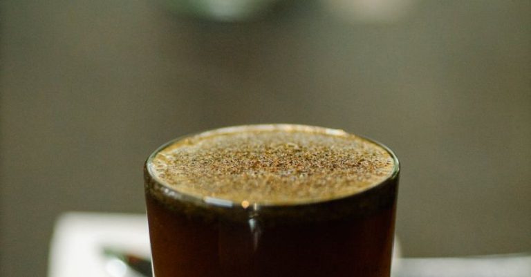 Coffee Cupping - Glass cup of freshly brewed coffee with froth placed on table with spoon and piece of paper with numbers on blurred background during cupping