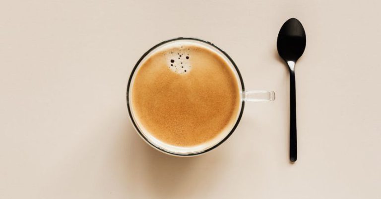 Coffee Blending - From above of clear glass cup with hot aromatic drink placed near black metal spoon on beige table for coffee break